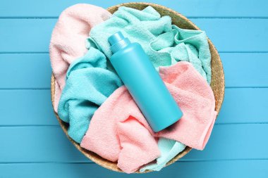 Açık mavi ahşap masa üzerinde havluyla çamaşır sepeti ve bir şişe deterjan.