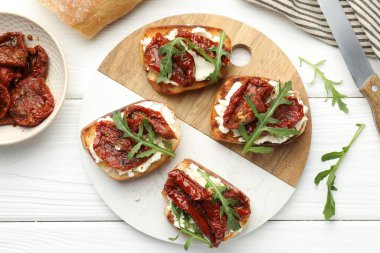 Krem peynirli nefis bruschettalar, güneşte kurutulmuş domatesler ve beyaz ahşap masada roka, düz uzanmış