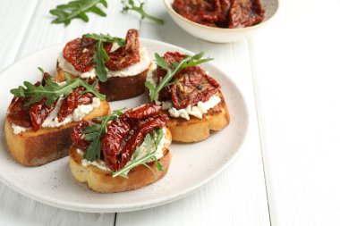 Krem peynirli lezzetli bruschettalar, güneşte kurutulmuş domatesler ve beyaz ahşap masada roka, yakın plan.