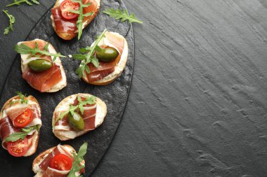 Delicious bruschettas with cream cheese, prosciutto, olives, tomatoes and arugula on dark textured table, top view. Space for text