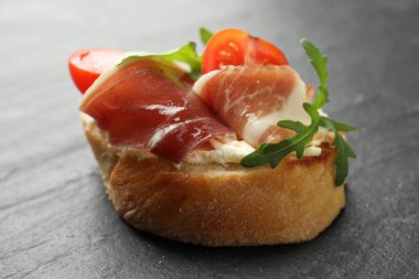 Delicious bruschetta with cream cheese, prosciutto, tomatoes and arugula on dark textured table, closeup