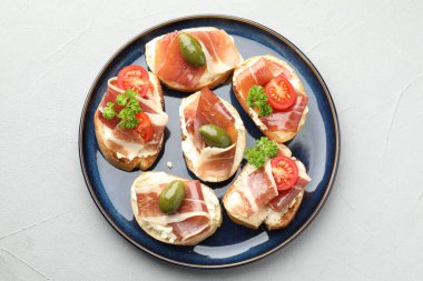 Delicious bruschettas with cream cheese, prosciutto, green olives, tomatoes and parsley on gray textured table, top view