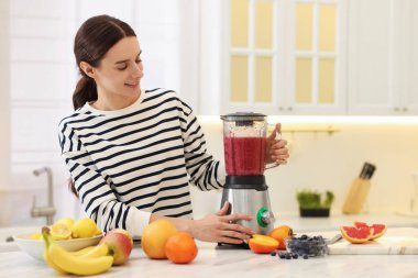 Gülümseyen kadın mutfaktaki beyaz mermer masada blender ile nefis smoothie yapıyor.