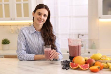Mutfaktaki beyaz mermer masada bir bardak lezzetli smoothie, blender ve meyvelerle gülümseyen kadın.