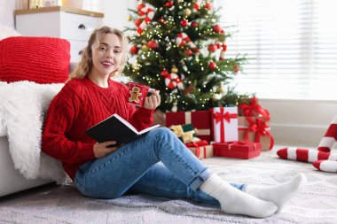 Süveter giyen, elinde bir bardak sıcak içecek ve kitapla gülümseyen kadın Noel için dekore edilmiş odada bulanık ışıklara karşı.