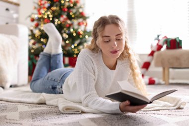 Süveter giymiş güzel bir kadın yerde kitap okuyor. Odadaki bulanık ışıklara karşı. Noel için süslenmiş.