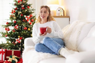Kanepede bir fincan sıcak içecekle süveter giyen gülümseyen kadın Noel için dekore edilmiş odadaki bulanık ışıklara karşı.