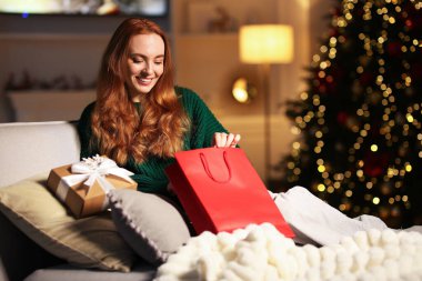 Kanepede Noel hediyeleri taşıyan süveterli bir kadın rahat bir odada bulanık ışıklara karşı. Metin için boşluk