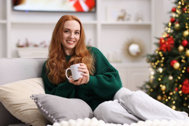 Sıcak bir bardak sıcak içecekle kanepede, rahat bir odada bulanık ışıklara karşı gülümseyen bir kadın. Mutlu Noeller ve mutlu yıllar.