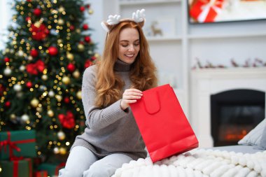 Evde bulanık ışıklara karşı elinde alışveriş çantasıyla ren geyiği saç bandıyla gülümseyen kadın. Mutlu Noeller ve mutlu yıllar.
