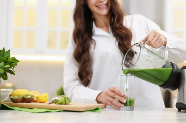 Gülümseyen kadın, beyaz masada, blenderdan cama lezzetli bir meyve püresi dolduruyor.