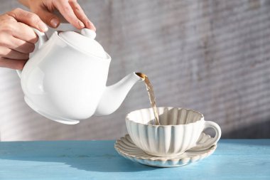 Açık mavi ahşap masada çaydanlıktan fincana sıcak çay dolduran kadın, yakın plan.