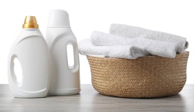 Çamaşır sepetinde havlular ve beyaz arka plandaki ahşap masada duran deterjan şişeleri.
