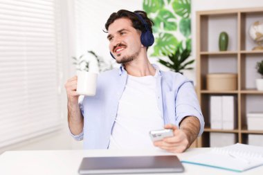 Kulaklıklı, akıllı telefonlu ve laptoplu gülümseyen adam, beyaz masada kapalı otururken bardaktan su içiyor.