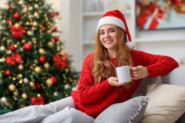 Noel Baba şapkalı, koltukta sıcak bir bardak içecekle gülümseyen kadın rahat odada bulanık ışıklara karşı, mesaj için yer var. Mutlu Noeller ve mutlu yıllar.