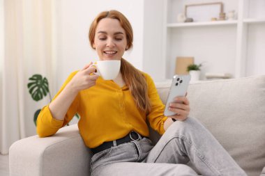 Evdeki kanepede bir fincan kahve ve akıllı telefonla güzel bir kadın.
