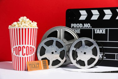 Kağıt bardakta lezzetli patlamış mısır, sinema bileti, alkış ve film makaraları beyaz masada kırmızı arka planda, yakın planda.