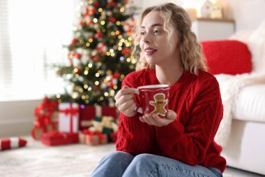 Süveter giymiş, elinde bir bardak sıcak içecekle gülümseyen bir kadın. Odadaki bulanık ışıklara karşı, Noel süslemesi yapılmış. Metin için boşluk