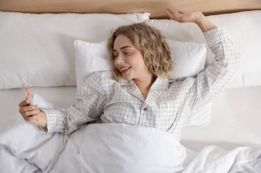 Akıllı telefonlu bir kadın evinde yatakta geriniyor.