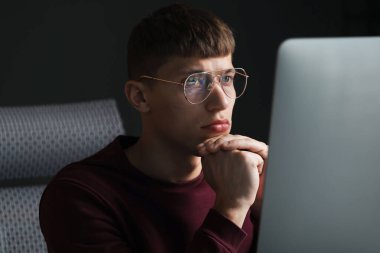 Kapalı alandaki bilgisayarın yanında yansıması olan gözlüklü genç adam.