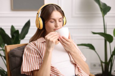 Elinde bir fincan aromatik kahve olan kadın evdeki koltukta müzik dinliyor.