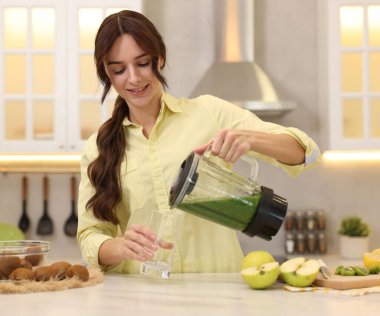 Gülümseyen kadın mutfaktaki beyaz mermer masada blenderdan cama lezzetli bir smoothie dolduruyor.