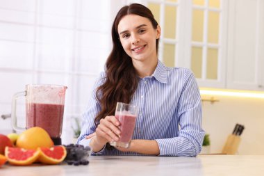 Mutfaktaki beyaz mermer masada bir bardak lezzetli smoothie, blender ve meyvelerle gülümseyen kadın.