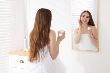 Havluya sarılı güzel bir kadın banyoda aynanın yanında kozmetik ürünler uyguluyor.