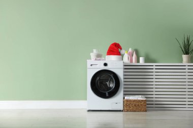 Noel Baba şapkalı çamaşır makinesi, çamaşır deterjanı ve sepet, odada adaçayı rengi yeşil duvarın yanında.