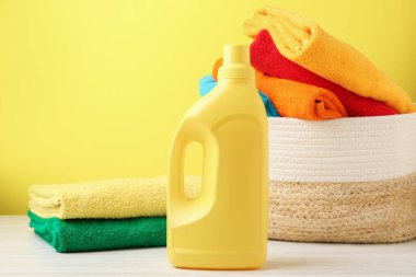Çamaşır sepetinde havlular ve beyaz ahşap masada bir şişe deterjan, sarı arka planda, yakın plan.