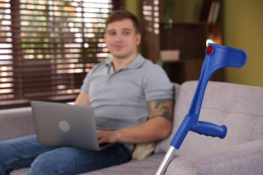 Evde dizüstü bilgisayar ve dirsek değneği üzerinde çalışan bir adam, seçici odaklanma