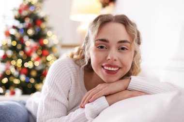 Evdeki bulanık ışıklara karşı kanepede süveter giymiş gülümseyen kadın portresi. Mutlu Noeller ve mutlu yıllar.