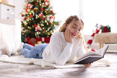 Süveter giyen, yerde kitap okuyan gülümseyen kadın Noel süslemeleri içinde bulanık ışıklara karşı.