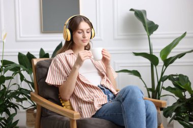 Elinde bir fincan aromatik kahve olan kadın evdeki koltukta müzik dinliyor.