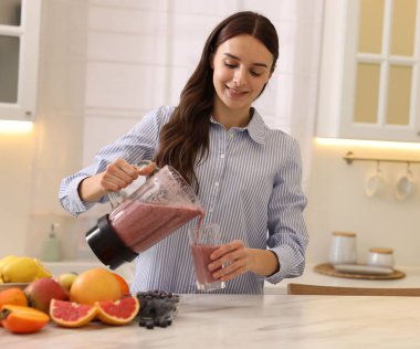 Gülümseyen kadın mutfaktaki beyaz mermer masada blenderdan cama lezzetli bir smoothie dolduruyor.