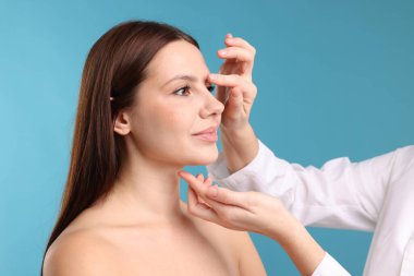 Açık mavi arka planda estetik ameliyattan önce hastanın yüzünü kontrol eden doktor, yakın plan.