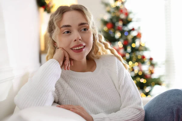 Evdeki bulanık ışıklara karşı kanepede süveter giyen gülümseyen kadın. Mutlu Noeller ve mutlu yıllar.