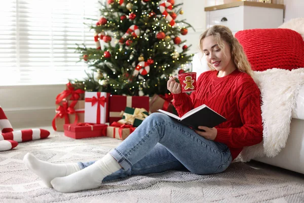 Süveter giymiş, elinde bir fincan sıcak içecek ile gülümseyen bir kadın yerde kitap okuyor Noel için dekore edilmiş odada bulanık ışıklara karşı.