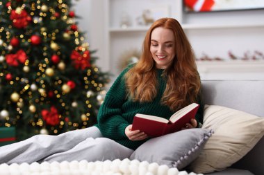 Sıcak odada bulanık ışıklara karşı kanepede kitap okuyan süveter giymiş gülümseyen kadın, mesaj için yer. Mutlu Noeller ve mutlu yıllar.