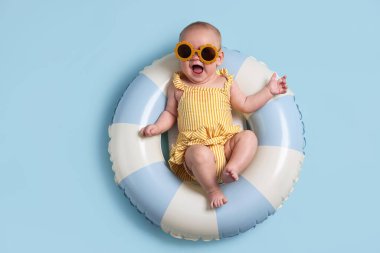 Şişirilebilir halkada güneş gözlüklü şirin bir bebek açık mavi arka planda, üst tarafta.