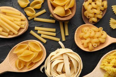 Tahta kaşıklardaki farklı çiğ makarna türleri siyah masada, düz yatıyordu.