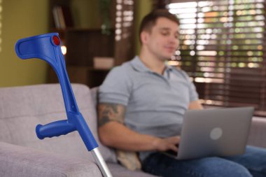 Evde dizüstü bilgisayar ve dirsek değneği üzerinde çalışan bir adam, seçici odaklanma