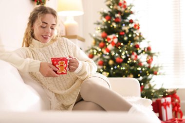 Süveter giymiş, elinde bir fincan sıcak içecekle gülümseyen kadın evdeki bulanık ışıklara karşı koltukta dinleniyor, mesaj için yer var. Mutlu Noeller ve mutlu yıllar.