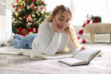 Süveter giyen, yerde kitap okuyan gülümseyen kadın Noel süslemeleri içinde bulanık ışıklara karşı.