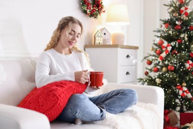 Kanepede bir fincan sıcak içecekle süveter giyen gülümseyen kadın Noel için dekore edilmiş odadaki bulanık ışıklara karşı.