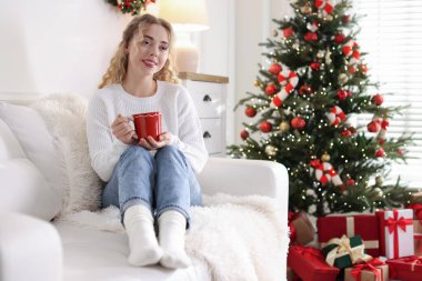 Koltukta bir fincan sıcak içecekle süveter giyen gülümseyen kadın Noel için dekore edilmiş odanın bulanık ışıklarına karşı. Metin için boşluk