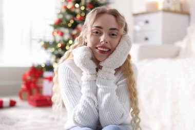 Evdeki bulanık ışıklara karşı eldivenli ve kazaklı gülümseyen kadın portresi. Mutlu Noeller ve mutlu yıllar.