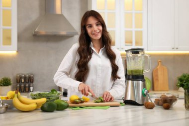 Gülümseyen kadın mutfakta blender ile beyaz mermer masada smoothie için kivi kesiyor.