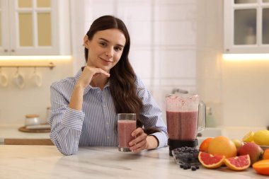 Mutfaktaki beyaz mermer masada bir bardak lezzetli smoothie, blender ve meyveli kadın.