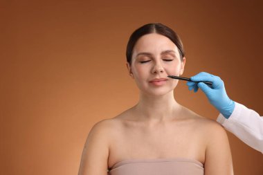 Doktor, kahverengi arkaplana karşı estetik ameliyat için hastanın yüzüne işaretler çiziyor, yakın plan. Metin için boşluk